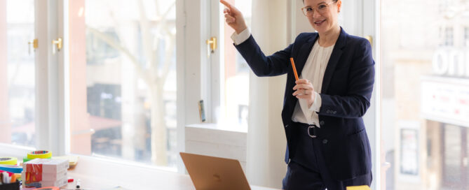 Salome Keller im Anzug mit Stift in der Hand vor Laptop und Moderationsmaterial, deutet nach vorn